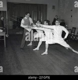 1960s, storici, studenti di sesso maschile che indossano tradizionali breches, giacche e pilmsols in una classe di scherma, con istruttore, Università di Oxford, Inghilterra, Regno Unito. Stanno lavorando alla spinta delle recinche, un movimento chiave nello sport, che è una spinta attaccante fatta con un piede in avanti e la gamba posteriore dritta e il braccio della spada allungato in avanti. Una vacanza sportiva su un tavolo sul retro della stanza, dice Roma 1960, un riferimento alle Olimpiadi di quell'anno. Foto Stock