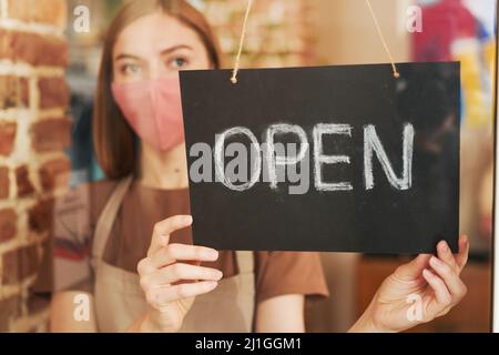 Orizzontale attraverso vetro porta ritratto di giovane cameriera indossare maschera protettiva sul viso di inizio giornata di lavoro con apertura caffè Foto Stock