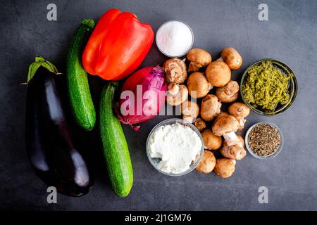 Insalata di verdure alla griglia ingredienti su sfondo scuro: Vista dall'alto di verdure crude, formaggio di capra, pesto e spezie Foto Stock