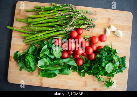 Vista dall'alto di verdure crude, erbe e spezie su un tagliere di legno: Pomodori ciliegini, asparagi, basilico, menta, prezzemolo, e spicchi d'aglio Foto Stock