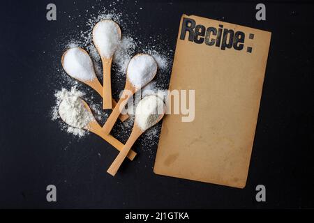 Ricetta stampata su una pagina di carta vintage su sfondo nero accanto a cucchiai di bambù con spezie bianche Foto Stock