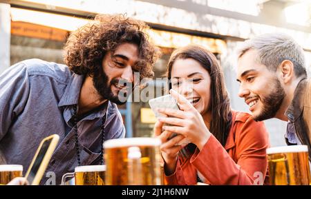 Giovani felici che si divertono con il telefono cellulare mentre bevono birra al bar birreria all'aperto - Focus sul viso della ragazza centrale Foto Stock