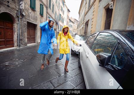 Una giovane coppia innamorata ha raggiunto un'auto mentre ha fatto una passeggiata in città in un clima allegro durante una giornata piovosa. Passeggiata, pioggia, città, relazione Foto Stock