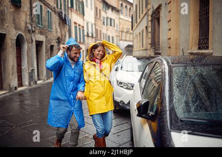 Una giovane coppia allegra in impermeabili viene catturata dalla pioggia durante una passeggiata in città in una giornata nuvolosa. Passeggiata, pioggia, città, rapporto Foto Stock