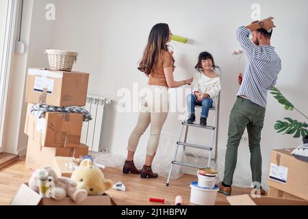 bambina seduta in scala tra i genitori in appartamento nuovo, giocando a dipingere il muro. famiglia giovane Foto Stock