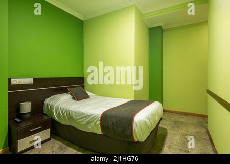 camera da letto con un piccolo letto di legno scuro con comodino coordinato, cuscino e coperta marrone con finiture rosse e pareti dipinte di verde pistacchio Foto Stock