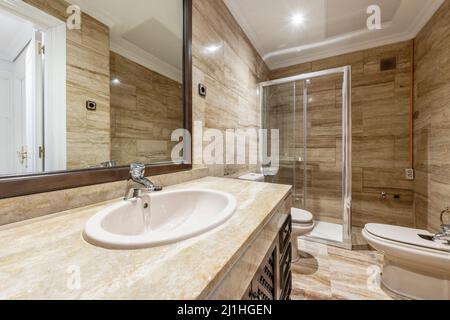 Bagno unico con lavandino in porcellana bianca con porte a rete su marmo color crema sotto specchio rettangolare con cornice marrone e cabina doccia Foto Stock