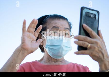 Gli anziani asiatici felici indossano una maschera facciale e rispondono a una videochiamata utilizzando il suo smartphone nel parco e chiacchierando con la famiglia, la tecnologia e il peo più vecchio Foto Stock