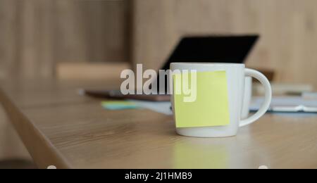 Una tazza di caffè bianca con una nota adesiva bianca gialla sulla tazza. Poggiato sulla scrivania e il computer portatile è posizionato sul retro. Foto Stock