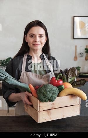Ritratto di sorridente donna attraente in grembiule che tiene in legno scatola piena di prodotti sani in cucina moderna con quadro astratto sul muro Foto Stock