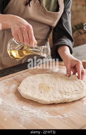 Primo piano di una mano femmina che versa l'olio dalla caraffa sull'impasto mentre si fa la pizza fatta in casa Foto Stock