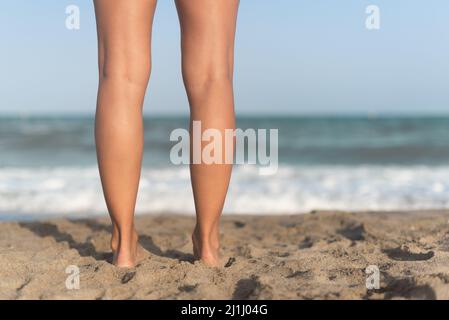 Gambe a piedi nudi femminili sulla spiaggia sabbiosa vicino al mare ondeggiante Foto Stock