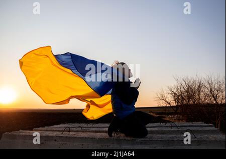 Silhouette di ragazzo in ginocchio, mani piegate in preghiera, appello a Dio. Dietro la schiena del bambino, la bandiera Ucraina fremola nel vento. Bambini contro la guerra. UKR Foto Stock