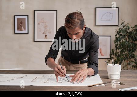 Concentrato il giovane ingegnere indiano di costruzione in camicia nera appoggiata sul tavolo e usando righello mentre lavora con Blueprint in ufficio Foto Stock