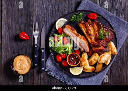 costolette di maiale alla griglia succose con spicchi di patate arrosto, lattuga fresca, pomodori su piatto nero, vista orizzontale, piatto Foto Stock