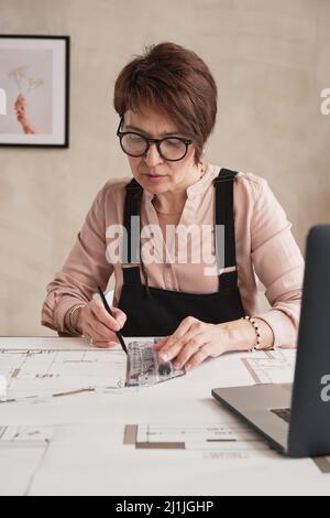 Donna matura concentrata in occhiali seduti al tavolo e usando righello mentre si fa piano pavimento in ufficio Foto Stock