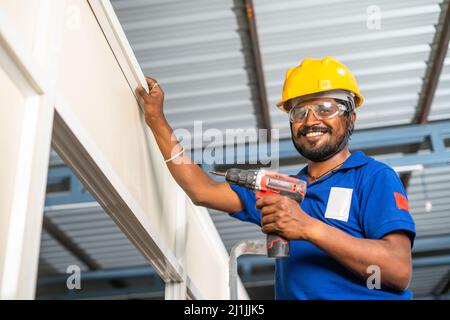 operatore che guarda la telecamera mentre fissa la vite utilizzando una macchina perforatrice o una pistola su un telaio divisorio in alluminio in cantiere - concetto di manutenzione Foto Stock