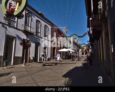 Vista sul centro storico della cittadina di Teror, Gran Canaria, Spagna con negozi e turisti che passano accanto e decorazioni natalizie in giornata di sole. Foto Stock