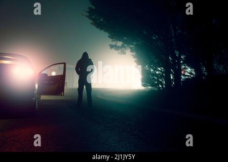 Un concetto oscuro di una figura minacciosa con cappuccio accanto a un'auto parcheggiata. In una strada cittadina di notte Foto Stock