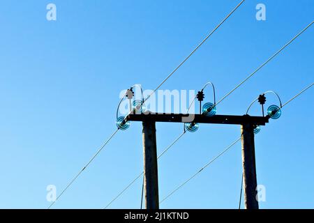 Un pilone rurale di legno di elettricità che trasporta tre linee di alimentazione ad alta tensione attraverso un campo, girato indietro illuminato contro un cielo blu luminoso. Foto Stock