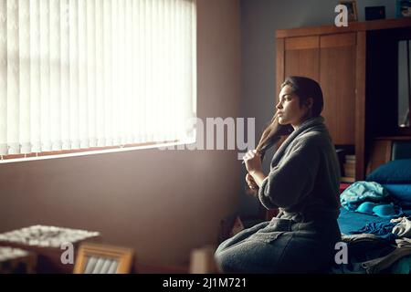 Pensando al giorno che sta davanti. Scatto di una giovane donna spazzolando i capelli mentre si siede sul suo letto la mattina. Foto Stock