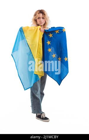 Orgogliosa e sconvolsa donna caucasica dai capelli biondi nel suo 20s tenendo insieme bandiere ucraine ed europee. Europa per l'Ucraina. Studio verticale girato su sfondo bianco. Foto di alta qualità Foto Stock