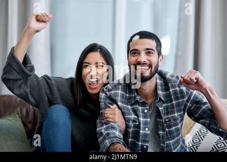 Chi ha bisogno del bar locale per divertirsi. Scatto di una giovane coppia felice che guarda la tv e che si rallegra a casa. Foto Stock