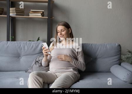 Sorridente giovane donna incinta usando il cellulare a casa. Foto Stock