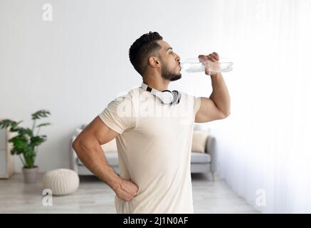Giovane uomo arabo sportivo con cuffie bere acqua dopo il suo allenamento a casa Foto Stock