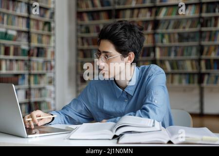 Studente ragazzo che studia, si siede al tavolo con il portatile in biblioteca Foto Stock