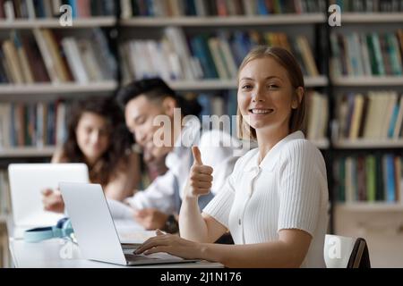 La studentessa si siede alla scrivania in biblioteca mostrando i pollici in su Foto Stock