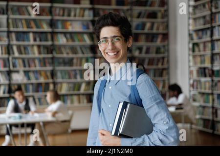 Lo studente tiene le cartelle di lavoro in posa in biblioteca, sorridendo guardando la fotocamera Foto Stock