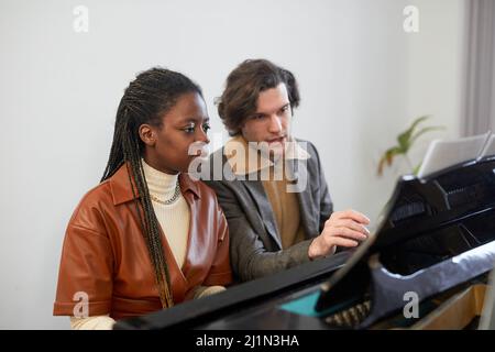 Giovane insegnante che punta alla spartiti e spiega alla donna come suonare il pianoforte durante la lezione musicale Foto Stock