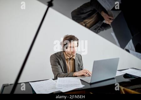 Studente serio che si concentra sul suo lavoro su un portatile mentre seduto al pianoforte grande, ha una lezione online Foto Stock