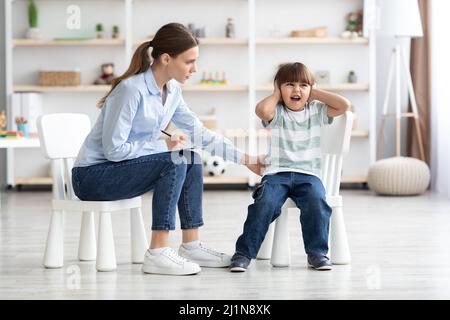 Ragazzo emozionale che urla e copre le orecchie durante la consultazione dello psicologo, donna che cerca di calmare il paziente Foto Stock