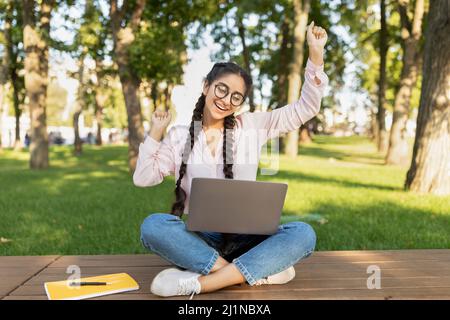 Studentessa indiana con un computer portatile che gesturava sì all'aperto, seduto nel campus universitario Foto Stock