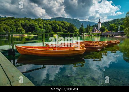 Ormeggiate barche a remi in legno sul lago e tradizionale chiesa alpina sullo sfondo, lago di Bohinj, Ribcev Laz, Slovenia, Europa Foto Stock