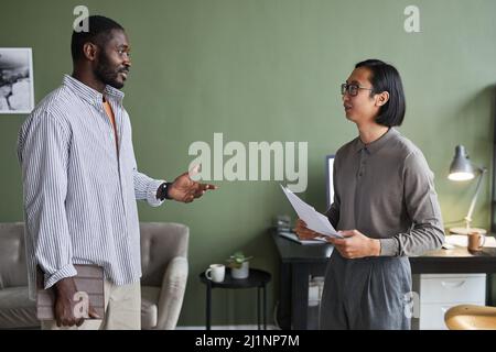 Ritratto laterale di due uomini d'affari etnici che discutono di lavoro mentre si trovano in un ufficio moderno contro il muro verde Foto Stock