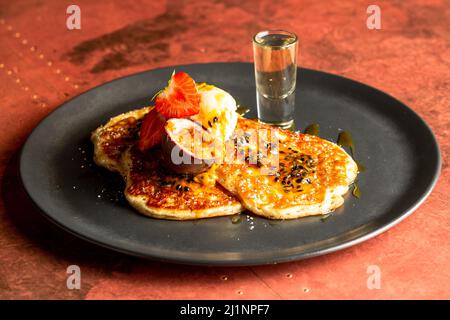 Un primo piano di pancake con fragole, frutto della passione e gelato servito su un piatto nero Foto Stock
