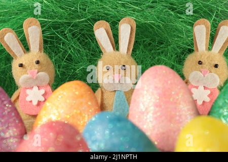 Tre coniglietti pasquali spaziati uniformemente separati dietro le uova pasquali colorate su uno sfondo verde e erboso. Foto Stock