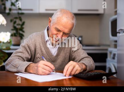 vecchio uomo grigio pelato siede a tavola e scrive su foglio bianco Foto Stock