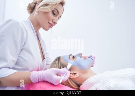 Beautician focalizzato applicazione maschera al volto di calma giovane donna Foto Stock