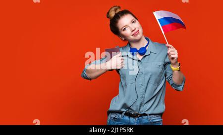 La studentessa caucasica ha una piccola bandiera russa e un passaporto in studio su sfondo arancione. Studiare all'estero concetto. Programma internazionale di scambio studentesco. Imparare il russo nel paese di origine. Foto Stock