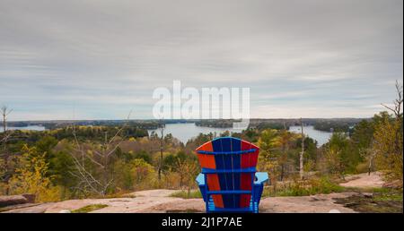 Una sedia Muskoka seduta su una roccia di fronte a una vista. Ci sono alcuni cottage accoccolati tra alberi verdi in condizioni di tempo calmo. Una serata nuvolosa. Messa a fuoco selettiva. Foto Stock