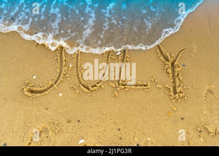 La parola luglio è scritta sulla sabbia di una spiaggia sul mare. Lavato via da un'onda. L'iscrizione scompare. Concetto di fine estate. Torna a scuola Foto Stock