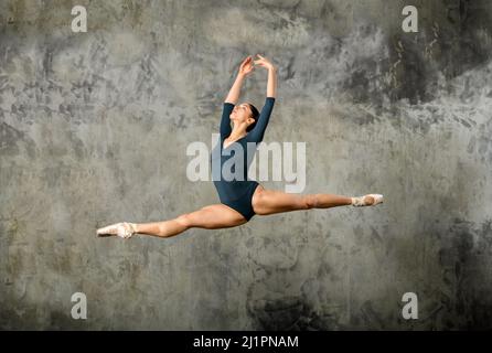 Corpo pieno di grazioso istruttore di balletto femminile in tuta e scarpe a punta che mostrano il classico salto gran jete contro il muro grigio di cemento Foto Stock