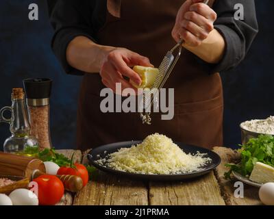 Cottura con formaggio. Lo chef strofina il formaggio su una grattugia manuale su un tavolo di legno su sfondo blu. Ingredienti per la cottura di pasta, pizza, torta, insalata. Clo Foto Stock