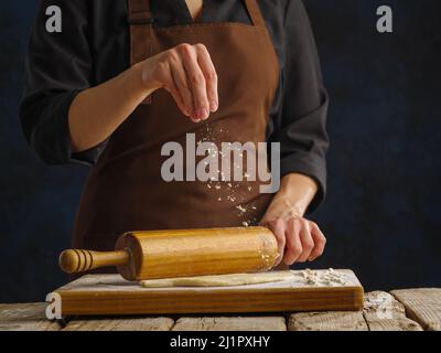 Lo chef stenderà l'impasto con un mattarello su un tagliere di legno su sfondo blu scuro. Primo piano. Concetto - ricette per pizza, torta, pasta Foto Stock