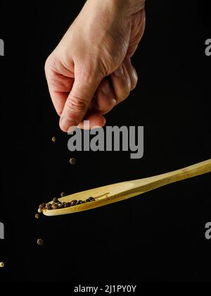 I piccoli grani di pepe in una mano della donna cadono su un cucchiaio di legno su uno sfondo nero. Levitazione. Minimalismo. Astrazione. Condimenti, spezie, ingredienti Foto Stock