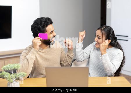 Allegra coppia indiana che guarda sullo schermo del computer portatile e sorride il toothy. Donna overjoyed gioire mentre facendo shopping in linea con suo marito. Trasferimento di denaro, concetto di online banking Foto Stock